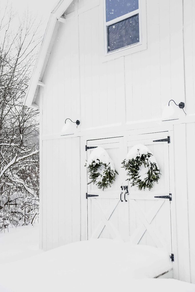 White barn with snowy farmhouse exterior and wreaths on doors and snow stacked on wreaths