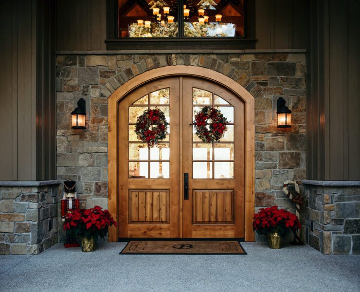 Wreaths hang from wood double doors framed by glass sconces and red flowers.