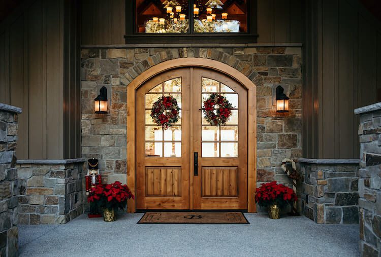 Wreaths hang from wood double doors framed by glass sconces and red flowers.