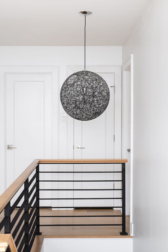 A mostly white interior with dark metal stairwell railing and a wood bannister in this global farmhouse style home. Above, a large light fixture meant to look like a woven basket ball hangs.