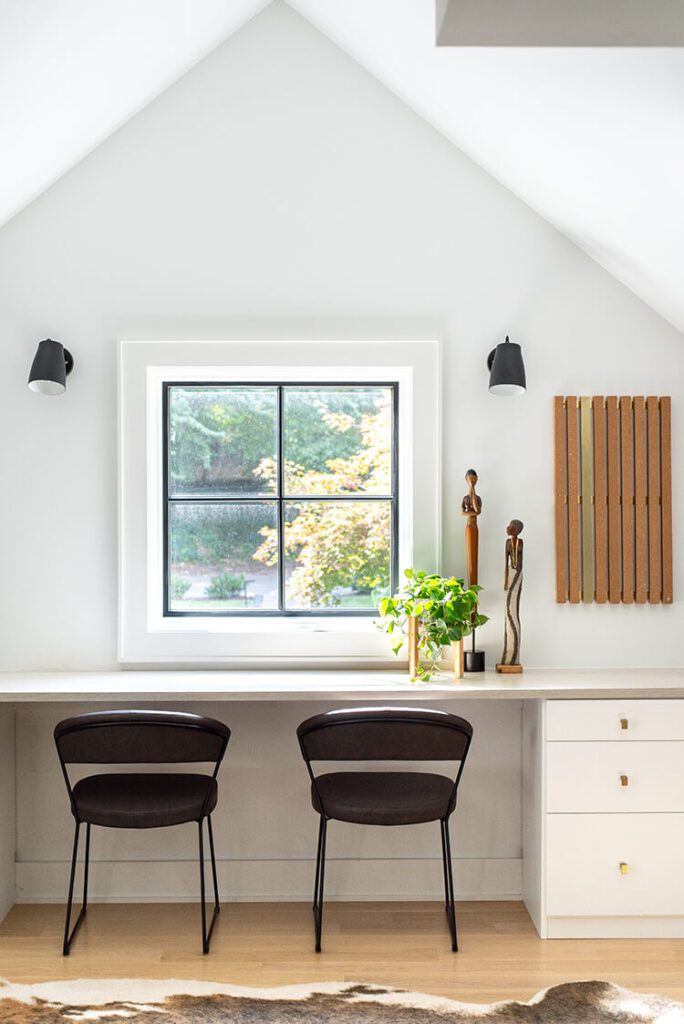 Inside this global farmhouse style home, a white interior has an A frame-like peak roof overlooking a square window with black metal farmhouse style window frames.