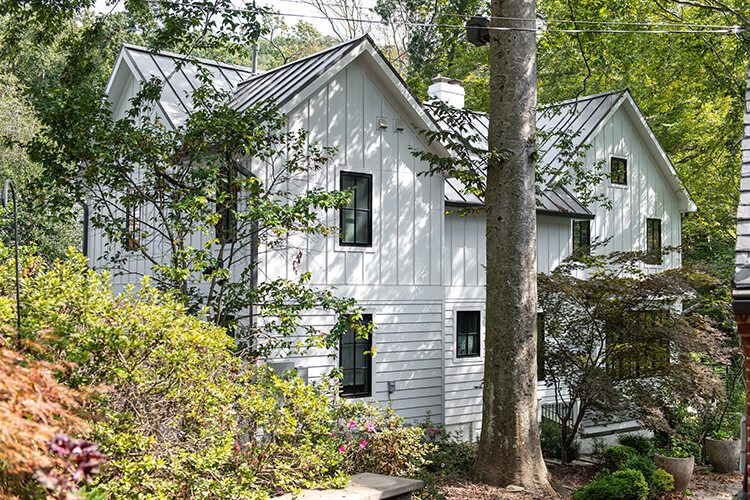 The exterior of a modern farmhouse style home with white paint is surrounded by verdant green trees.
