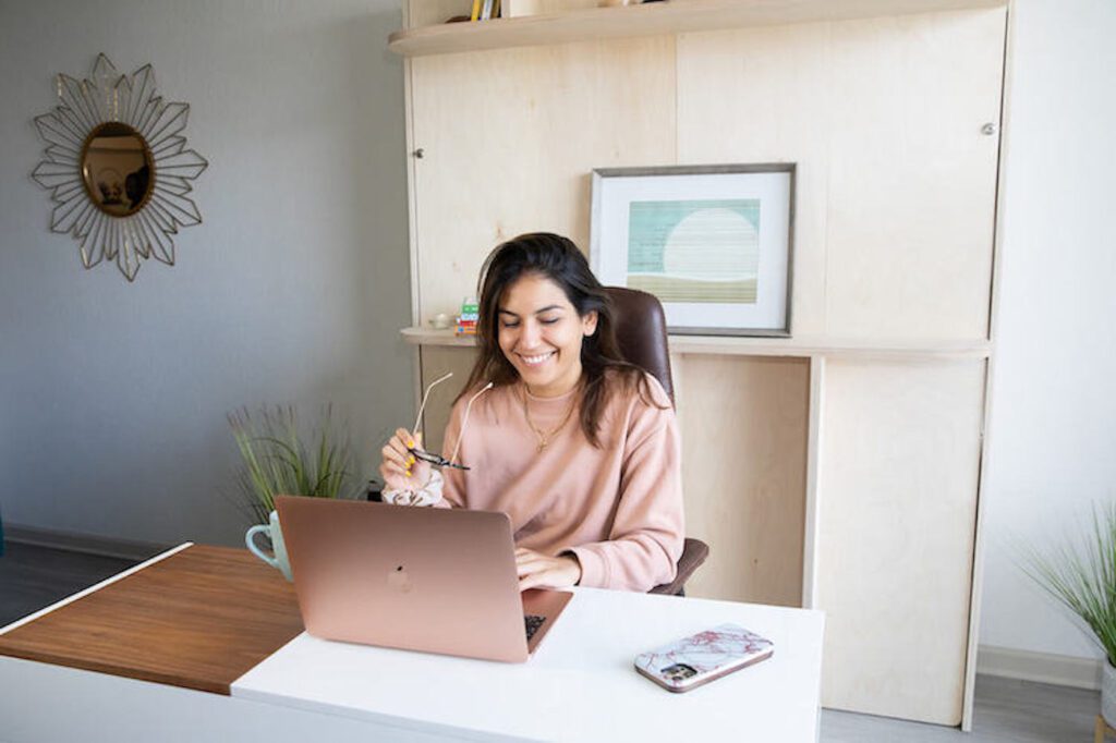A woman sits at her desk with a rose gold laptop and a Murphy bed in the background.