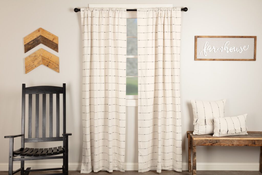 From left to right along a wall are a rocking chair and wood chevron art piece, window with cream drapery, “farmhouse” sign, and wood bench with two white throw pillows.