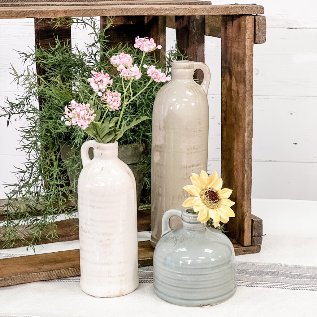 In front of a shiplap wall is a dark wood crate and two tall and one round ceramic jars holding flowers and greenery.