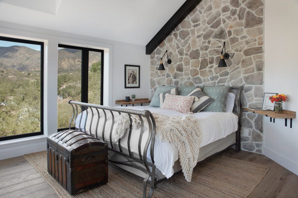 A stone wall acts as an accent wall behind the bed. Reclaimed vintage oak floors and little custom extras like the bedside shelf tables come together to give this space an open and airy feeling yet one which is warm and welcoming.