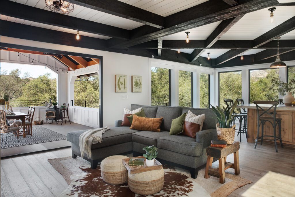 A gray counch sits under a beam ceiling made of dark wood. White shiplap covers some of the walls and the wood floor appears aged to make a warm farmhouse setting.