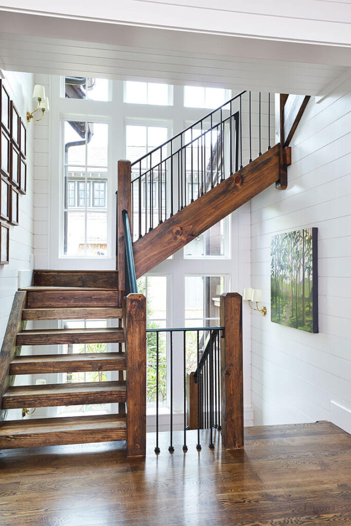 A wood staircase runs along a wall of windows descending up to the second story. The room is otherwise all white and shiplap.