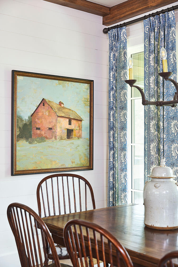 An oil painting of a horse stable offers subtle references to Georgian countryside. The painting overlooks a dark wood farmhouse table and hanging blue curtains in the window.