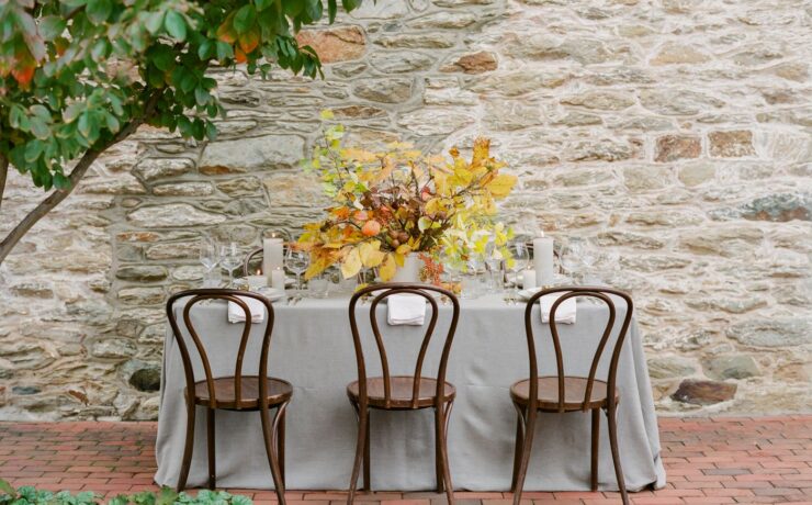 A white tablecloth covers a table outside near a stone wall. On the tablecloth sits tall white pillar candles, a centerpiece filled with gold and yellow fall leaves, and white plates with a small french horn