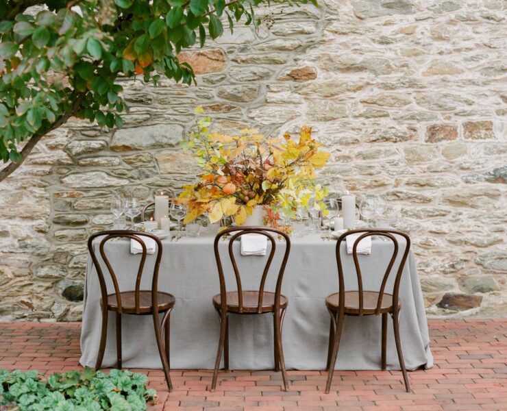 A white tablecloth covers a table outside near a stone wall. On the tablecloth sits tall white pillar candles, a centerpiece filled with gold and yellow fall leaves, and white plates with a small french horn