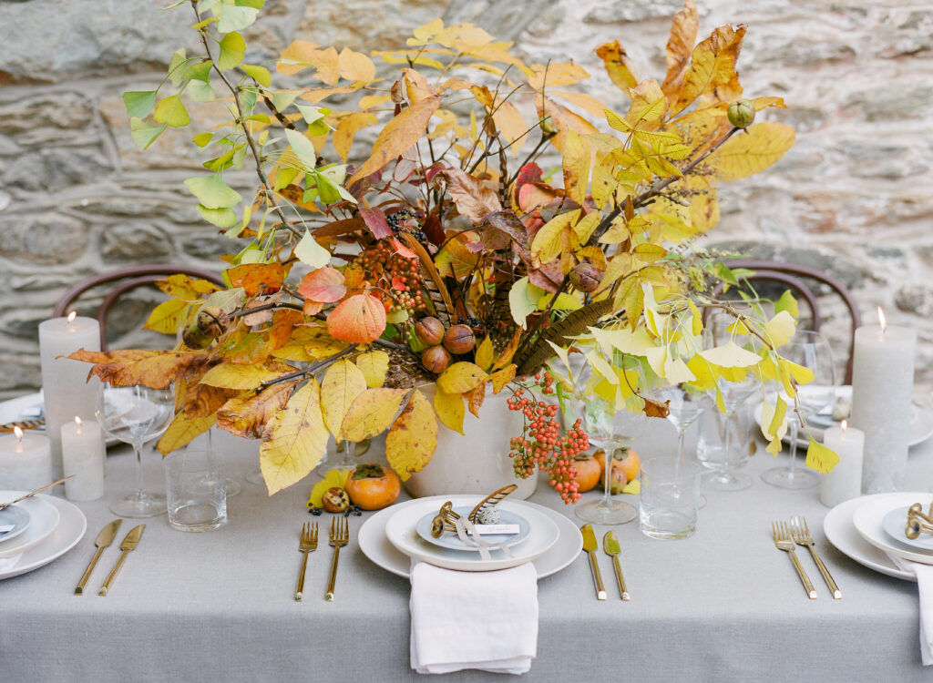 A white tablecloth covers a table outside near a stone wall. On the tablecloth sits tall white pillar candles, a centerpiece filled with gold and yellow fall leaves, and white plates with a small french horn