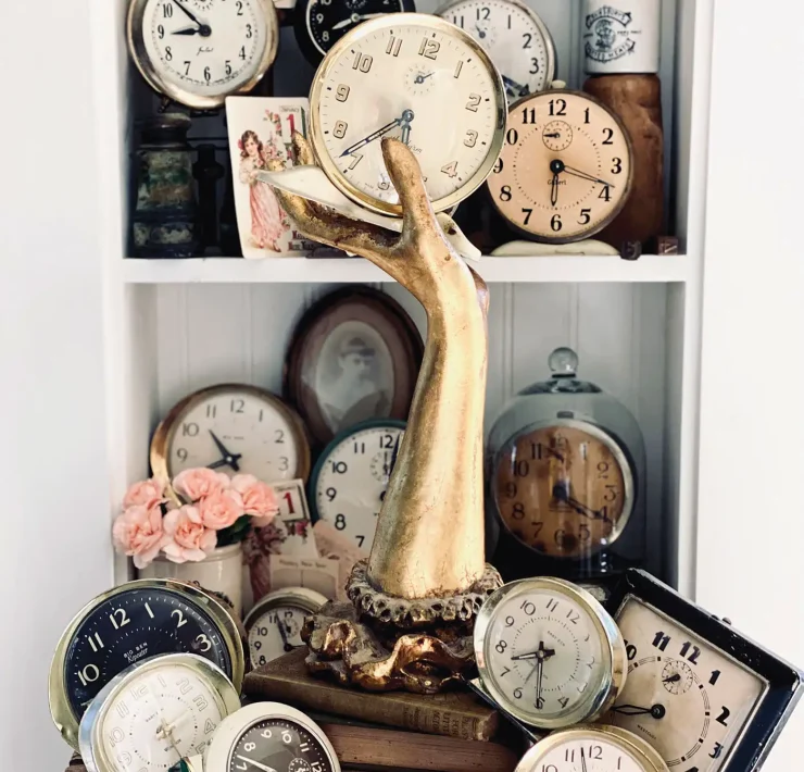 collection of vintage clocks arranged on a bookshelf with brass hand and other antiques