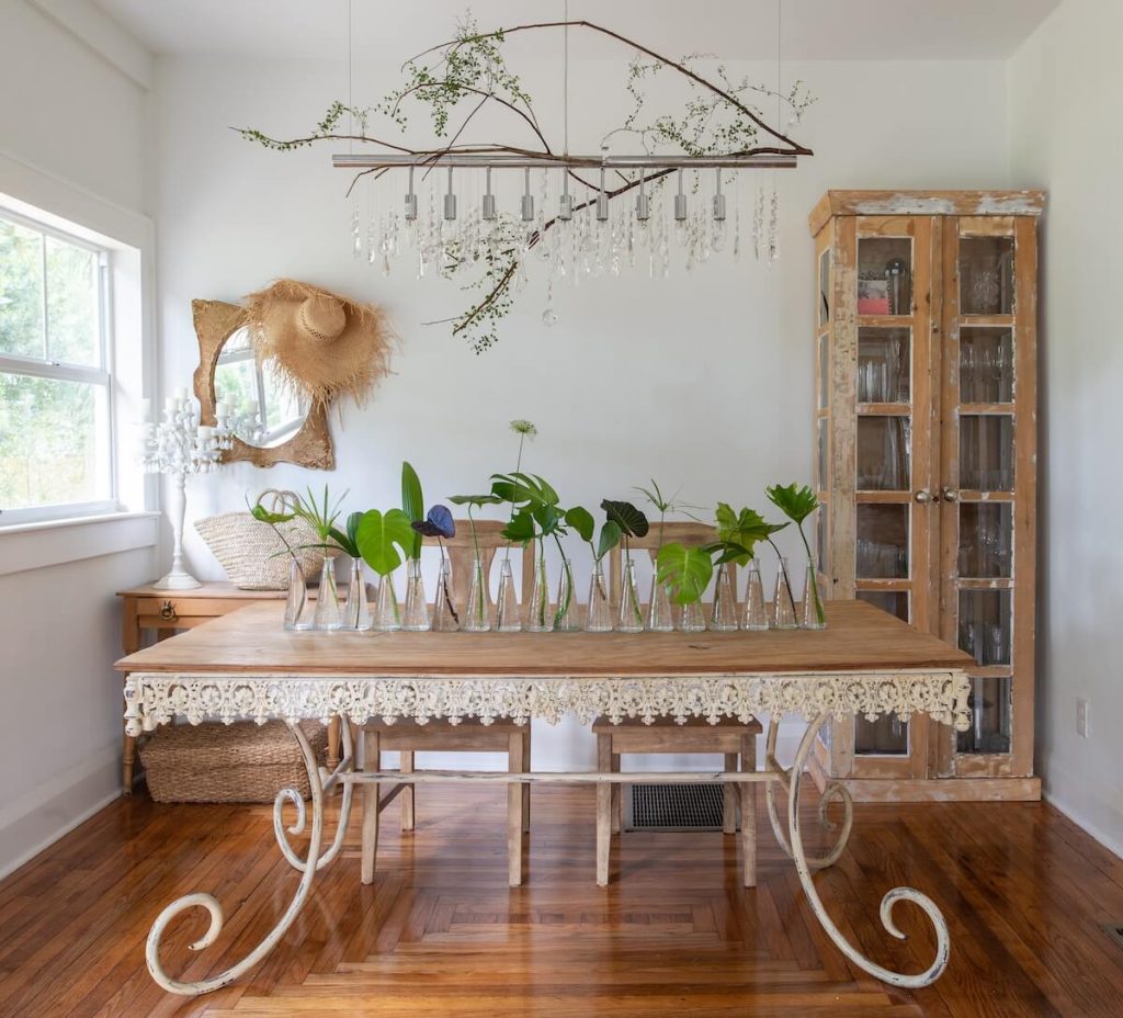 chandelier with tree branch hanging on it and collection of bud vases with monstera and palm leaves on antique table below